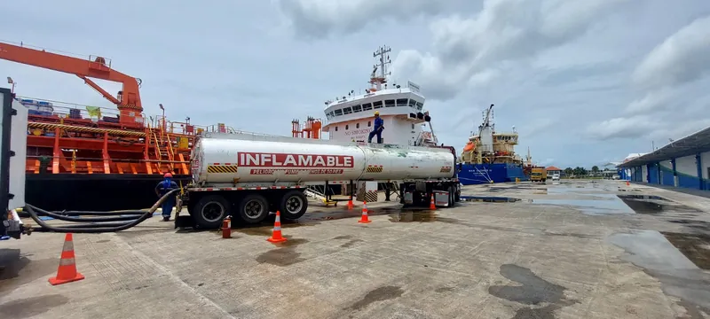 Tank truck marked “Inflamable” connected by hose to a tanker ship on a wet port apron