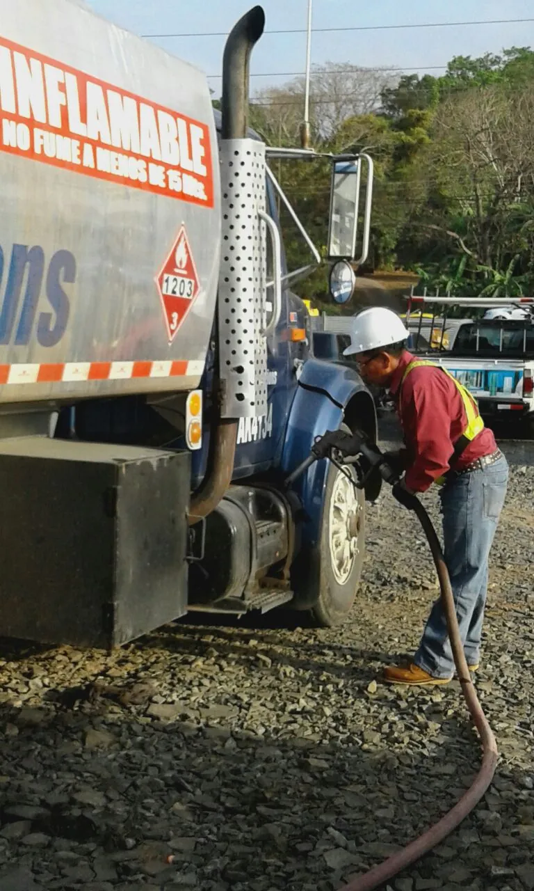 Operador con casco blanco y chaleco reflectivo sosteniendo la pistola de una manguera de combustible junto a un camión cisterna
