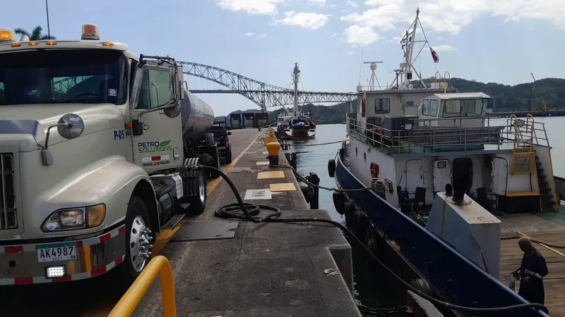 PetroSolutions tank truck on a pier next to a moored vessel, with a steel arch bridge in the background
